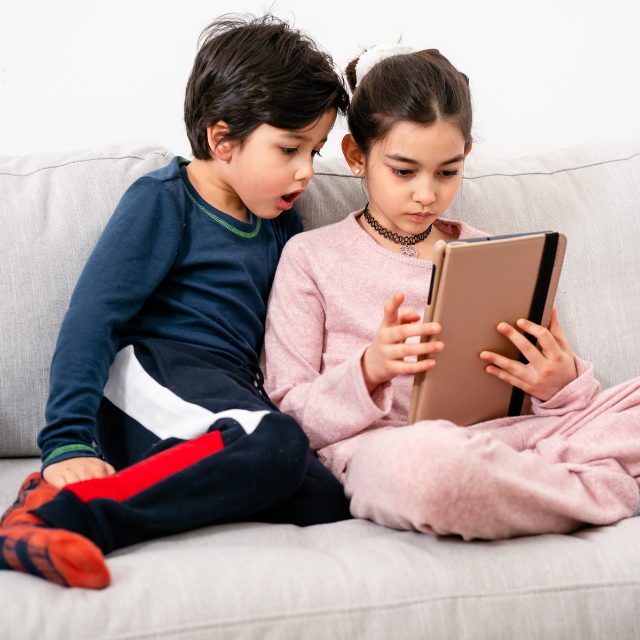 Tow children sitting on a grey couch watching an iPad